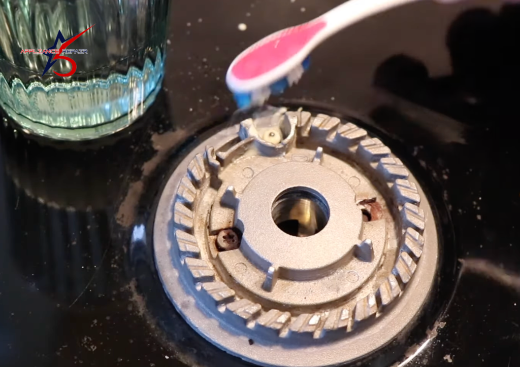 A close-up showing a person using a pink toothbrush to clean food debris and grease from a gas stove burner head and spark igniter.