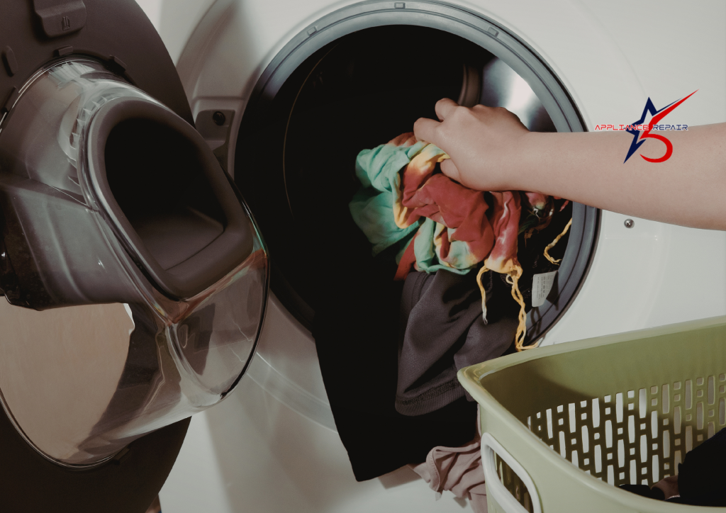 person’s hand moving a load of colorful laundry from a green plastic basket into a white front-load clothes dryer