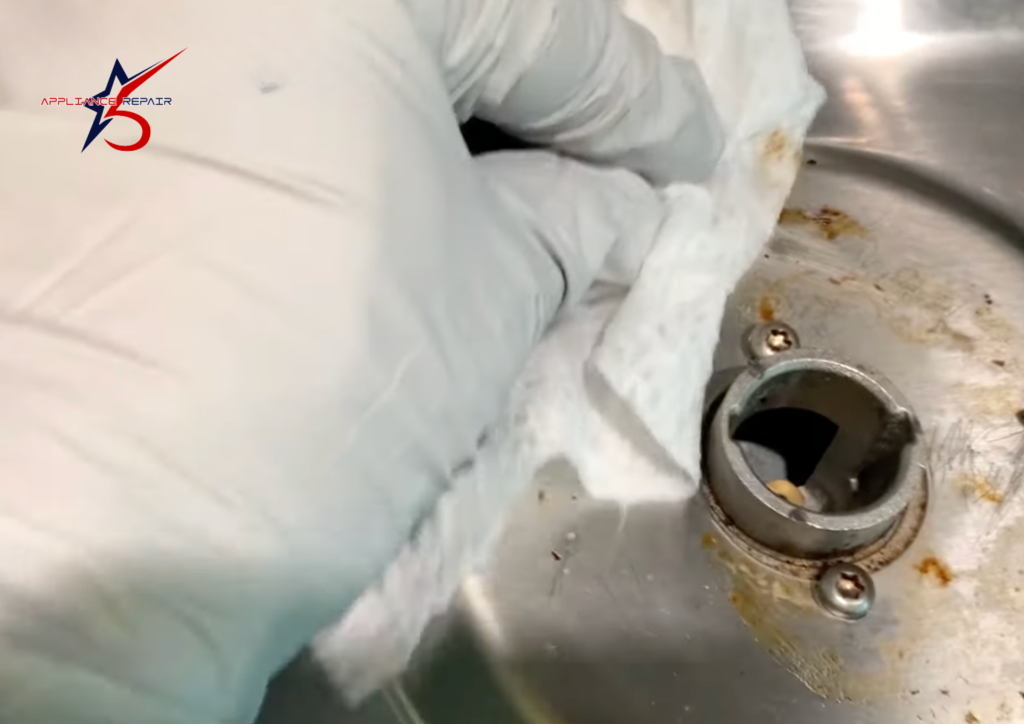 A gloved technician using a white cloth to wipe away grease and residue from a stainless steel gas stove burner base.