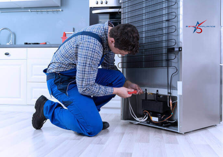 A 5 Star Appliance technician in blue overalls kneeling to repair the compressor and condenser coils on the back of a stainless steel refrigerator in a modern kitchen.