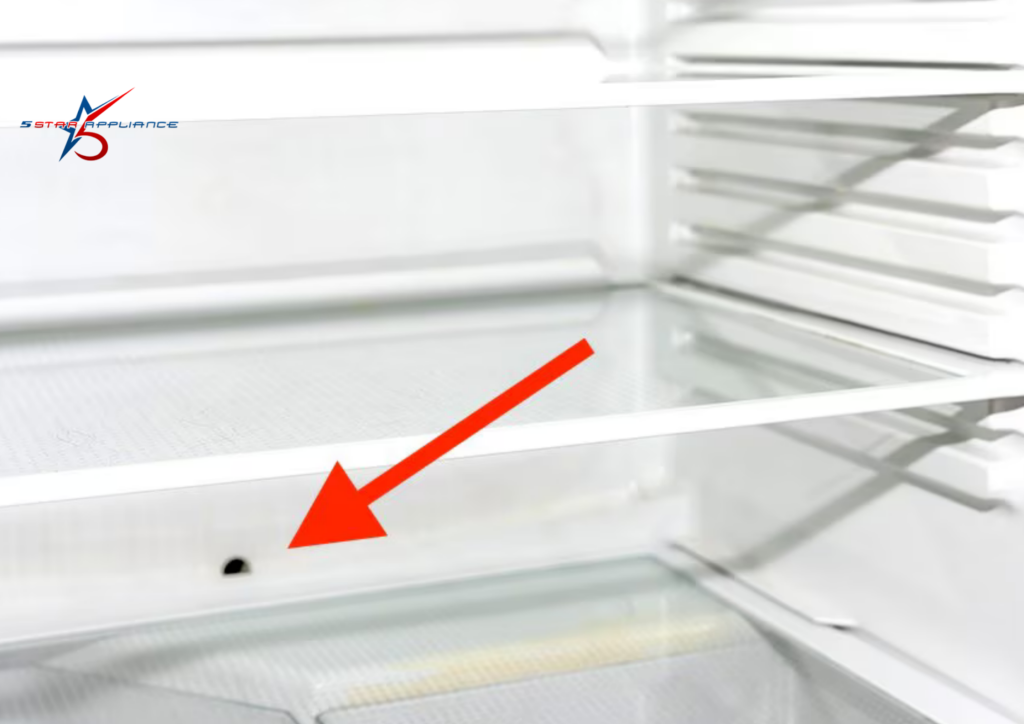 A close-up of the interior back wall of a white refrigerator with a red arrow pointing to a small defrost drain hole located just above the glass vegetable crisper shelf.