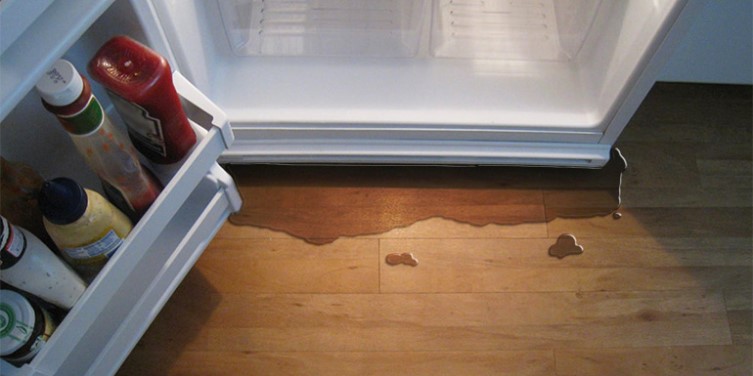 A kitchen refrigerator leaking water onto a hardwood floor, requiring troubleshooting and repair.